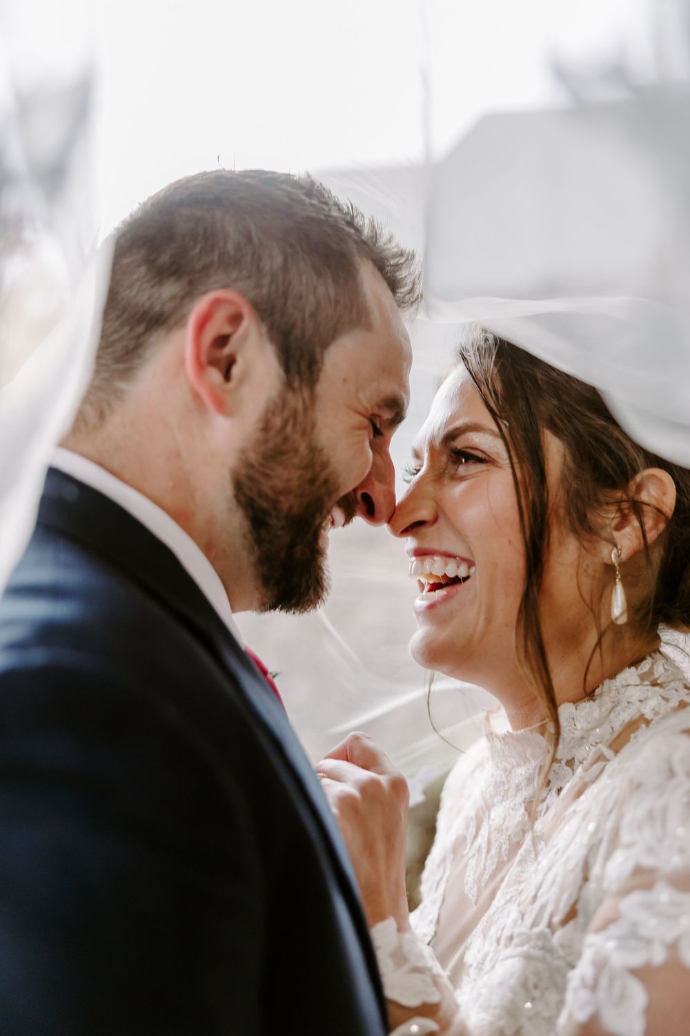 Bride and groom share an intimate moment under a veil, smiling and touching foreheads on their wedding day.