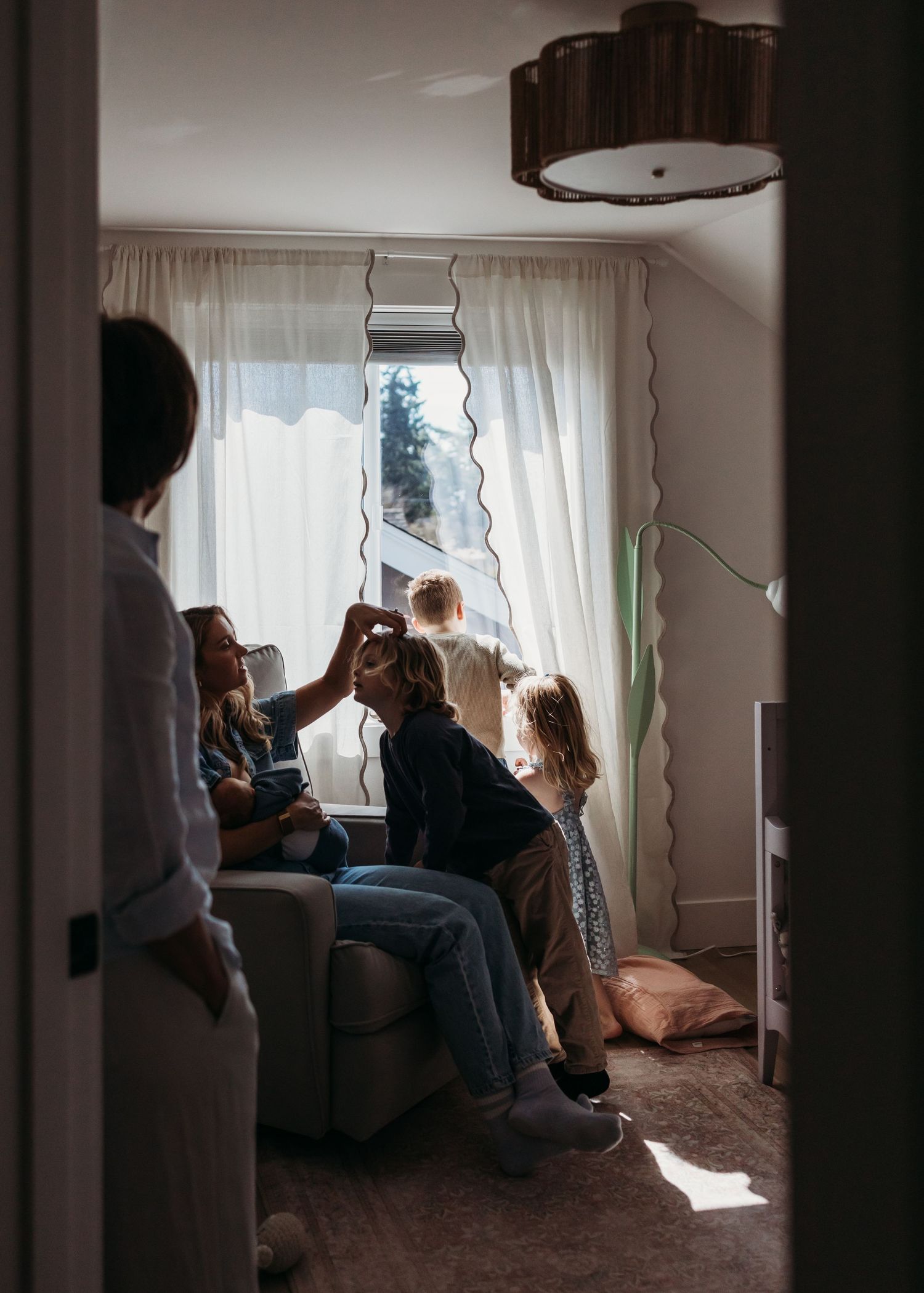 Family gathered in a cozy living room by a sunlit window, with children playing and adults sitting on a sofa.