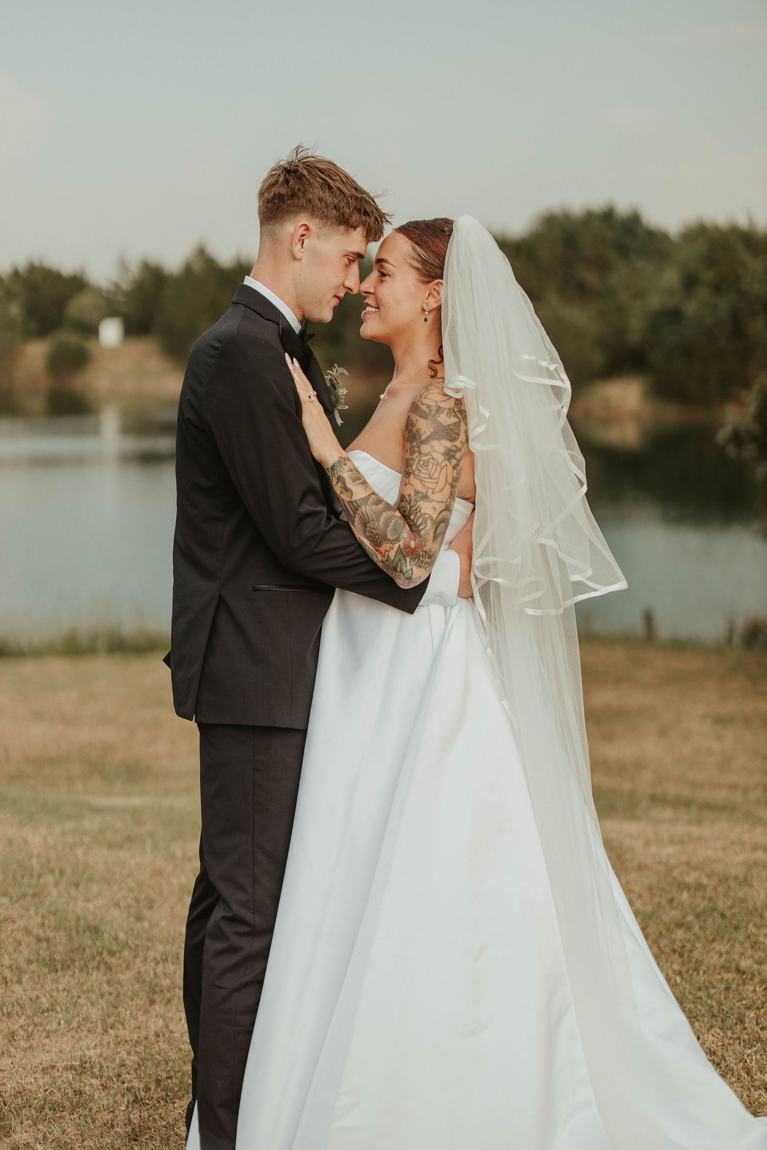 Bride in white gown and veil with groom in black tuxedo sharing romantic moment outdoors near lake.