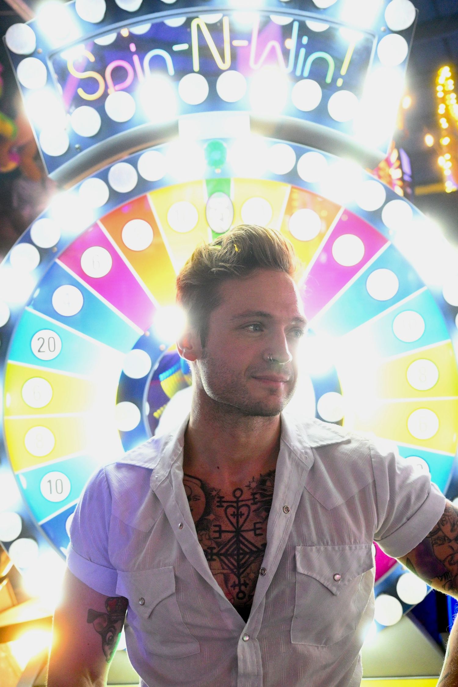 Tattooed man standing in front of a colorful illuminated Spin-N-Win carnival game wheel at a fair.