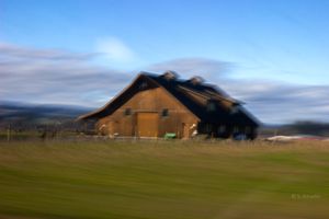 Motion blur photograph of a rustic brown barn in a green field under a blue sky.