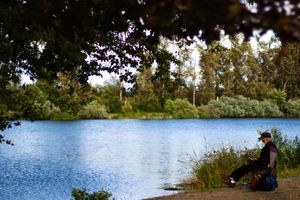 Person sitting by a calm blue lake surrounded by lush green trees on a cloudy day.