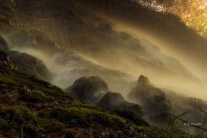 Misty waterfall cascading over moss-covered rocks with golden sunlight filtering through the haze in a lush forest.