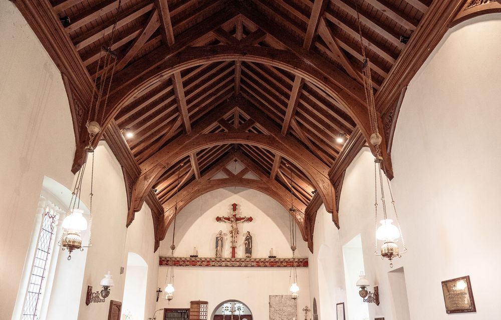 Gothic church interior with vaulted wooden ceiling, arched beams, crucifix altar, and pendant lights in a white-walled nave.