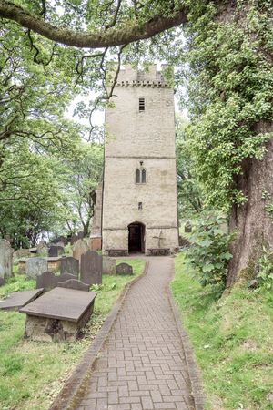 A stone church tower with battlements surrounded by trees and gravestones in an English churchyard.