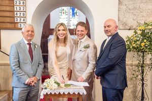 A bride and groom pose with two men beside a floral arrangement inside a church.