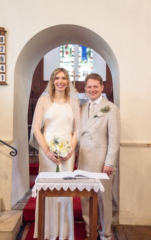 Bride and groom smiling together at church altar, bride in white gown with veil holding yellow bouquet, groom in beige suit.