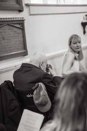 Elderly man in wheelchair at a wedding ceremony inside a historic church, with a blonde woman seated nearby.
