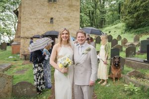 Bride and groom smiling in a churchyard with gravestones, guests with umbrellas, and a dog in the background.