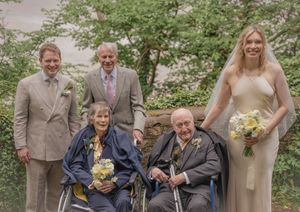 Wedding group photo with bride in ivory gown, groom in tan suit, and elderly guests in wheelchairs holding yellow bouquets.
