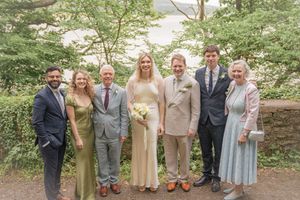 Wedding party of seven smiling adults posing outdoors among lush green trees, bride holding white bouquet in elegant setting.