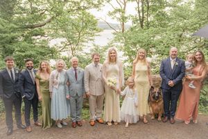 Wedding party poses outdoors among lush green trees, bride in white gown holding bouquet with family and dog.