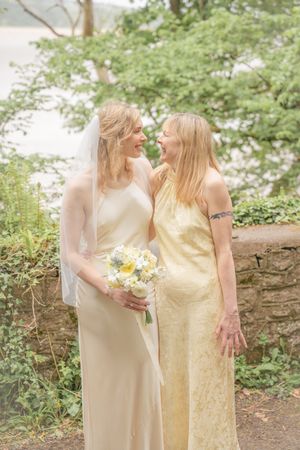 Two women in yellow dresses, one a bride with veil and bouquet, share a joyful moment outdoors.