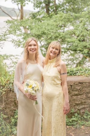 Two women in elegant cream and gold dresses pose together at an outdoor wedding ceremony.