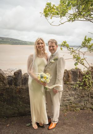 A bride and groom pose by a stone wall with a sandy estuary in the background.