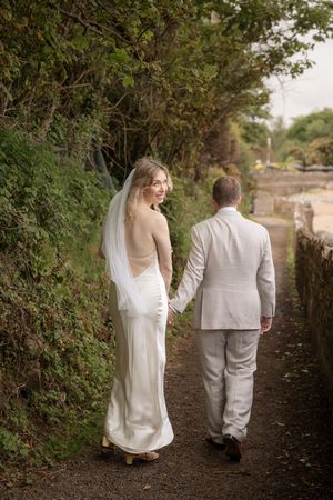 Bride in backless silk gown and veil holds grooms hand walking along tree-lined path after wedding.