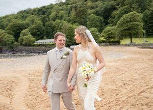 Bride and groom holding hands walking along a sandy beach with green hills behind them.