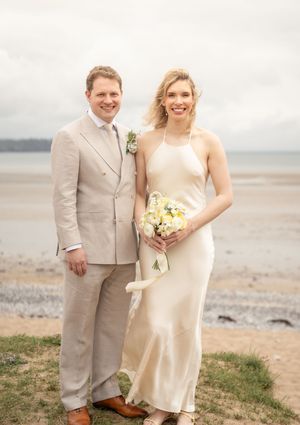 Bride and groom pose together on a sandy beach, she in a white halter dress with veil, he in a beige suit.