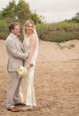 Happy bride and groom posing on sandy beach, bride holding white bouquet in elegant gown with veil.