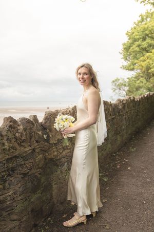 Blonde bride in ivory satin gown holding yellow and white bouquet poses by stone wall near seaside.