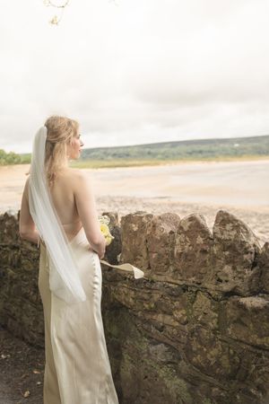 Bride in white satin gown and veil stands by stone wall overlooking a scenic beach landscape.