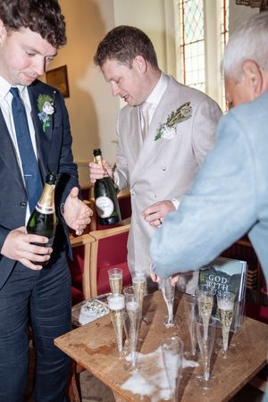 Two groomsmen in suits pouring champagne into flutes at a wooden table inside a church during a wedding reception.
