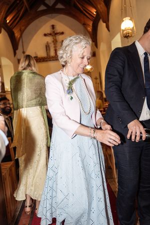 Elegant older woman in light blue dress and pink jacket smiling in a church during a wedding ceremony.
