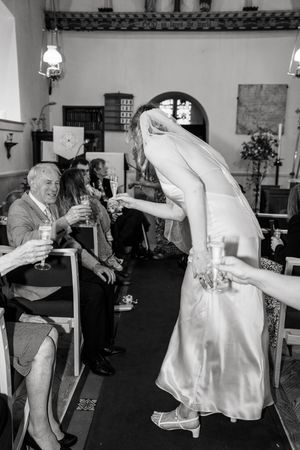 Bride in white gown and veil bends to greet seated wedding guests holding champagne glasses during ceremony.