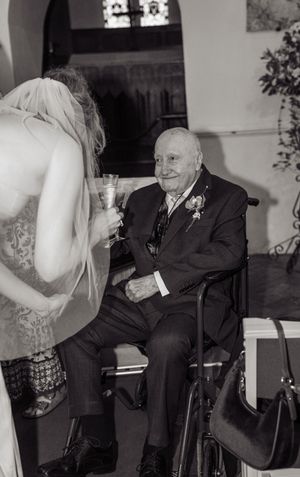 Black and white photo of a bride in a veil toasting with an elderly man in a wheelchair at a wedding ceremony.