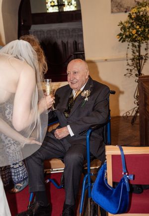 A bride in a veil toasts with an elderly man in a wheelchair at a church wedding.