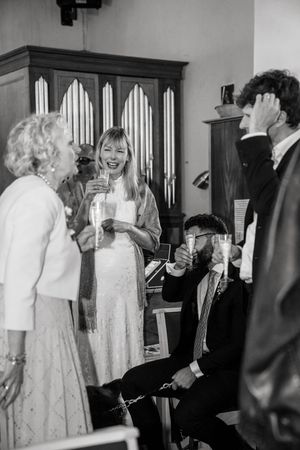Black and white photo of wedding guests laughing and toasting with champagne glasses at a reception.