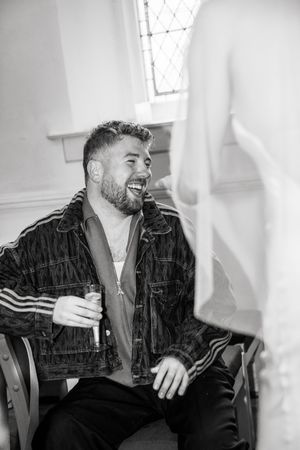 Black and white photo of a laughing bearded man in a denim jacket holding a drink while seated indoors.