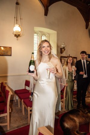 Smiling bride in white satin dress holding champagne bottle and glass inside a church.