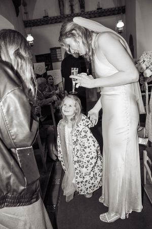 Bride in white satin gown bending down to interact with a smiling young girl at a wedding reception.