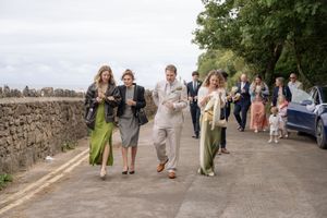 Wedding guests walking along a stone wall path, dressed in elegant attire including green dresses and a linen suit.