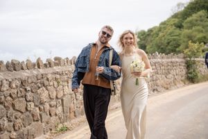 Couple walking on a country road, groom in denim jacket, bride in white dress holding bouquet, stone wall background.