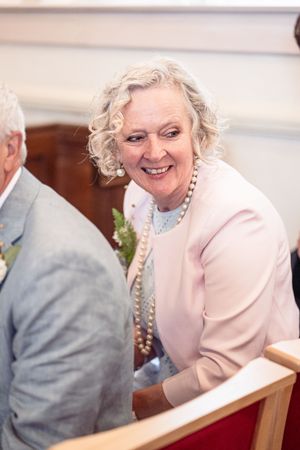 Elegant older woman with curly white hair and pearl necklace smiling warmly while seated in a church pew at a wedding.