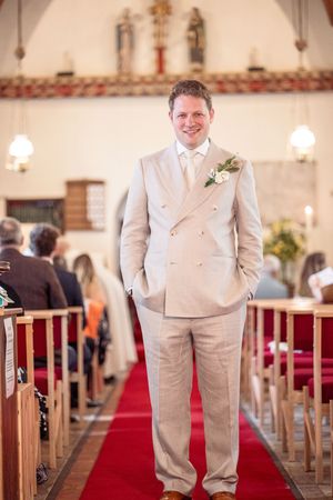 Groom in beige double-breasted suit with boutonniere stands at church altar aisle before wedding ceremony.
