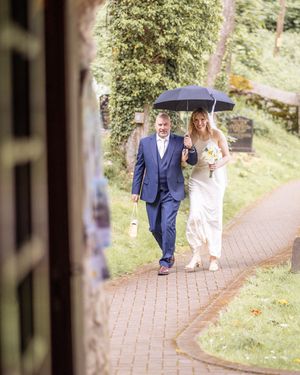 Bride and father walking to church under umbrella on wedding day.