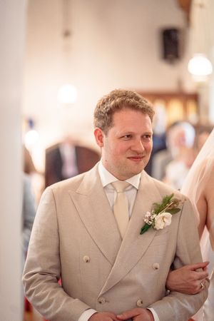 Smiling groom in tan double-breasted suit with floral boutonniere during wedding ceremony in church.