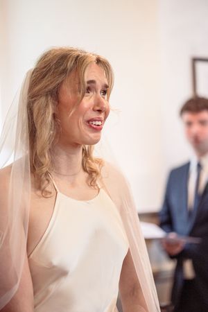 Emotional bride in white halter gown and veil smiles tearfully during wedding ceremony indoors.