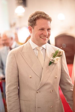 Smiling groom in beige double-breasted suit with floral boutonniere standing in church aisle on wedding day.