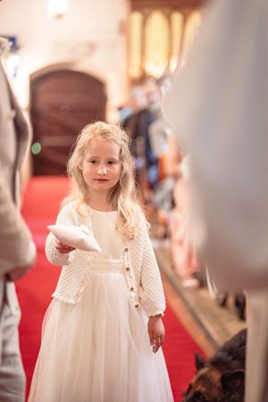 Young flower girl in white dress carrying a ring pillow down a red-carpeted church aisle.