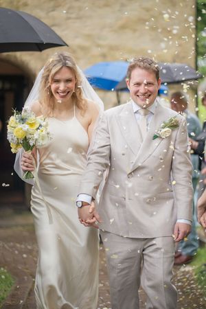 Newlyweds walking hand in hand through confetti shower, bride in white gown, groom in beige suit, under umbrella.