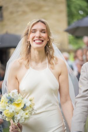 Radiant bride in sleeveless white gown and veil smiles joyfully while holding a yellow and white floral bouquet outdoors.
