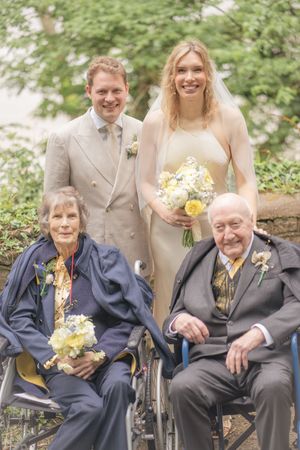 Wedding couple in cream attire poses with elderly grandparents in wheelchairs, holding yellow and white floral bouquets outdoors.