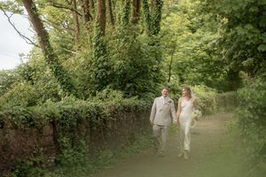 Newlywed couple walking hand in hand along lush green countryside path.