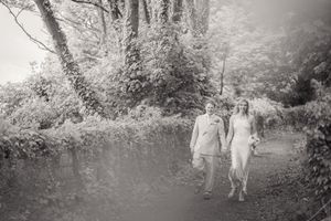 A black and white photo of a bride and groom walking along a tree-lined woodland path.