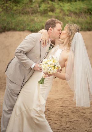 Bride and groom share a romantic kiss on sandy beach, bride holding yellow and white bouquet.