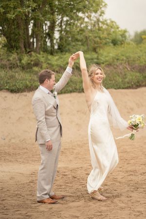 Groom in beige suit twirls smiling bride in white gown and veil on a sandy beach while she holds a bouquet.